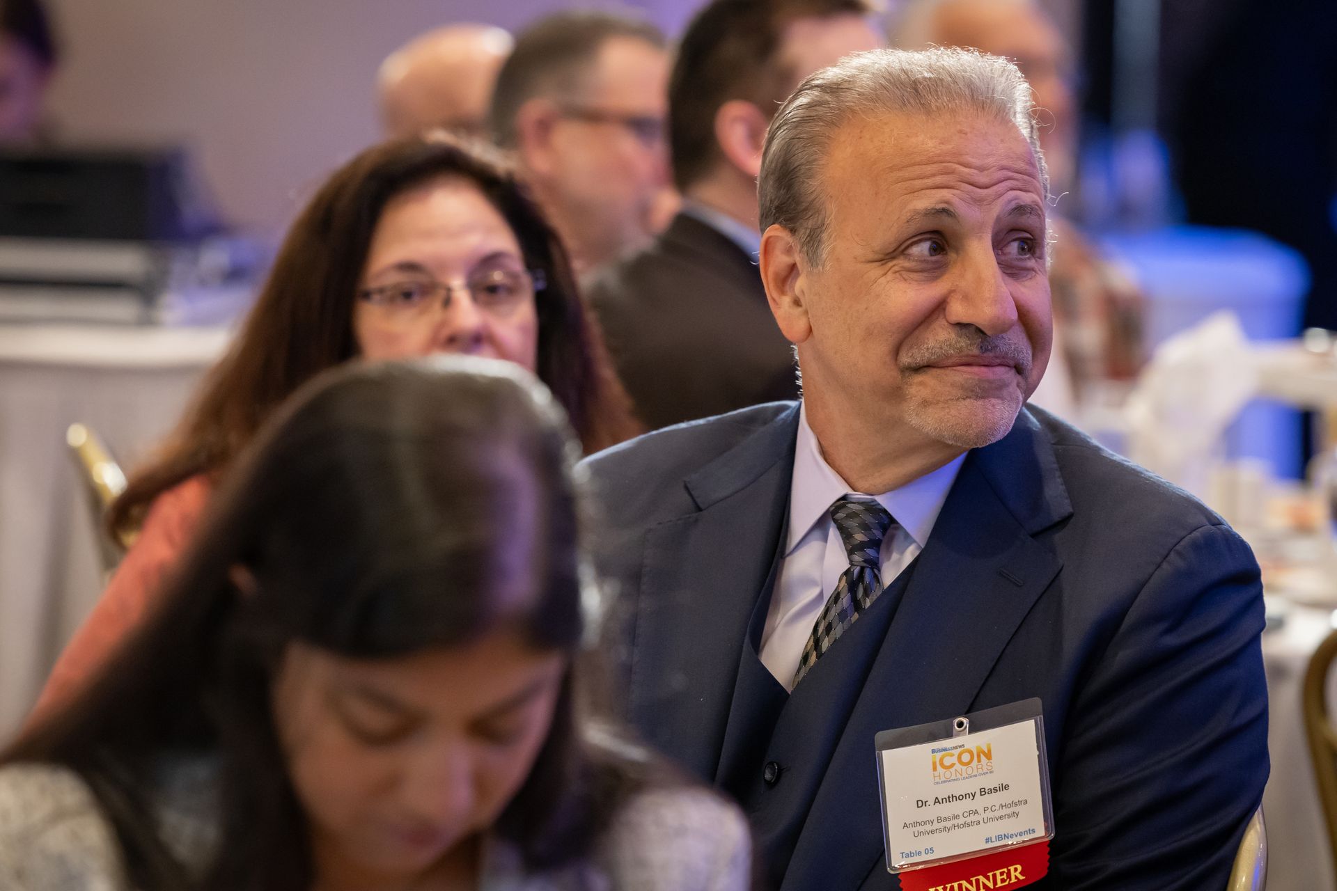 A candid close-up of Dr. Anthony Basile wearing a dark suit and name tag, seated at a table during an awards event.