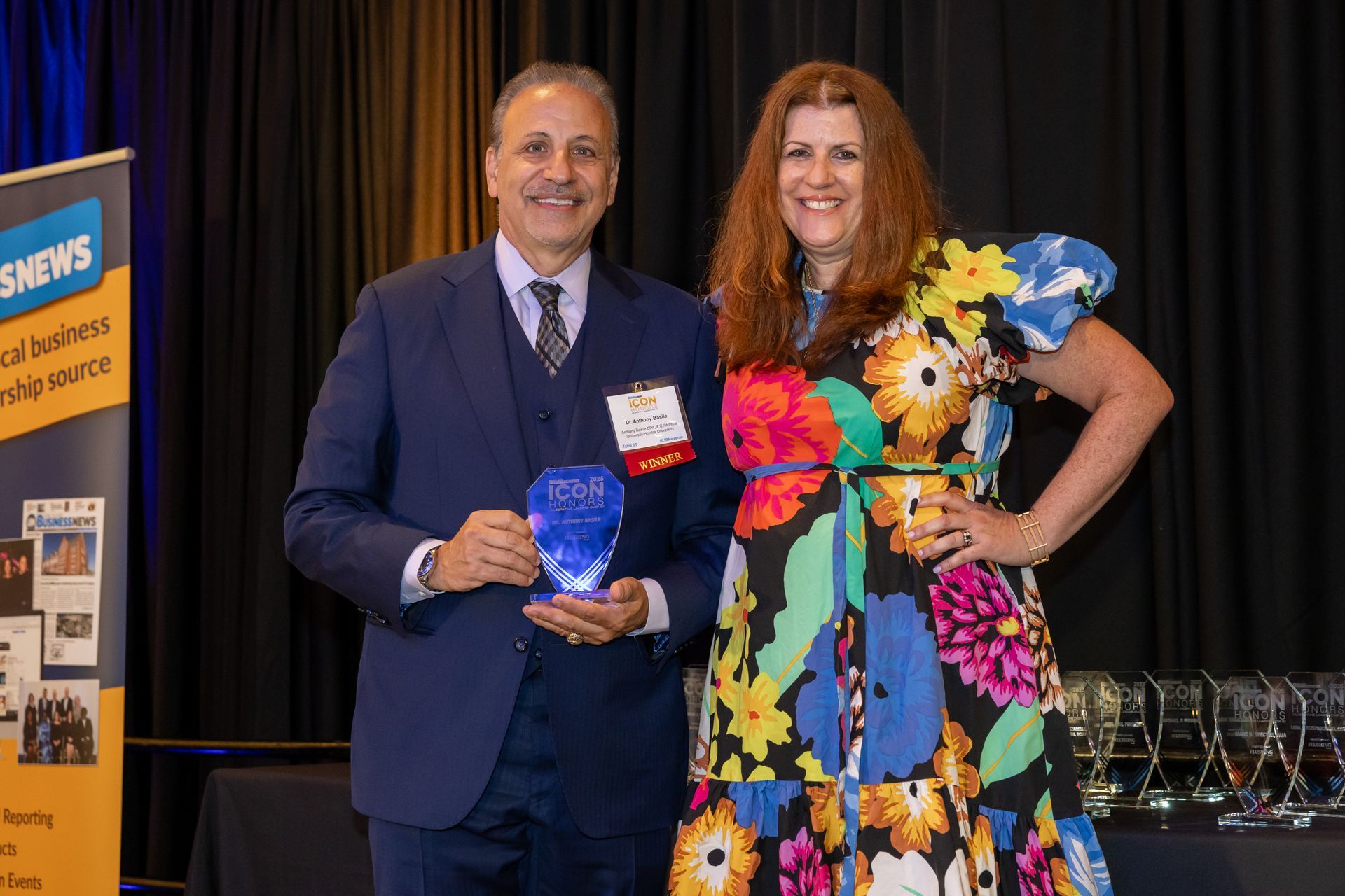 Dr. Anthony Basile holding a glass Icon Honors award trophy while posing alongside a woman in a colorful floral dress in front of a dark curtain backdrop.